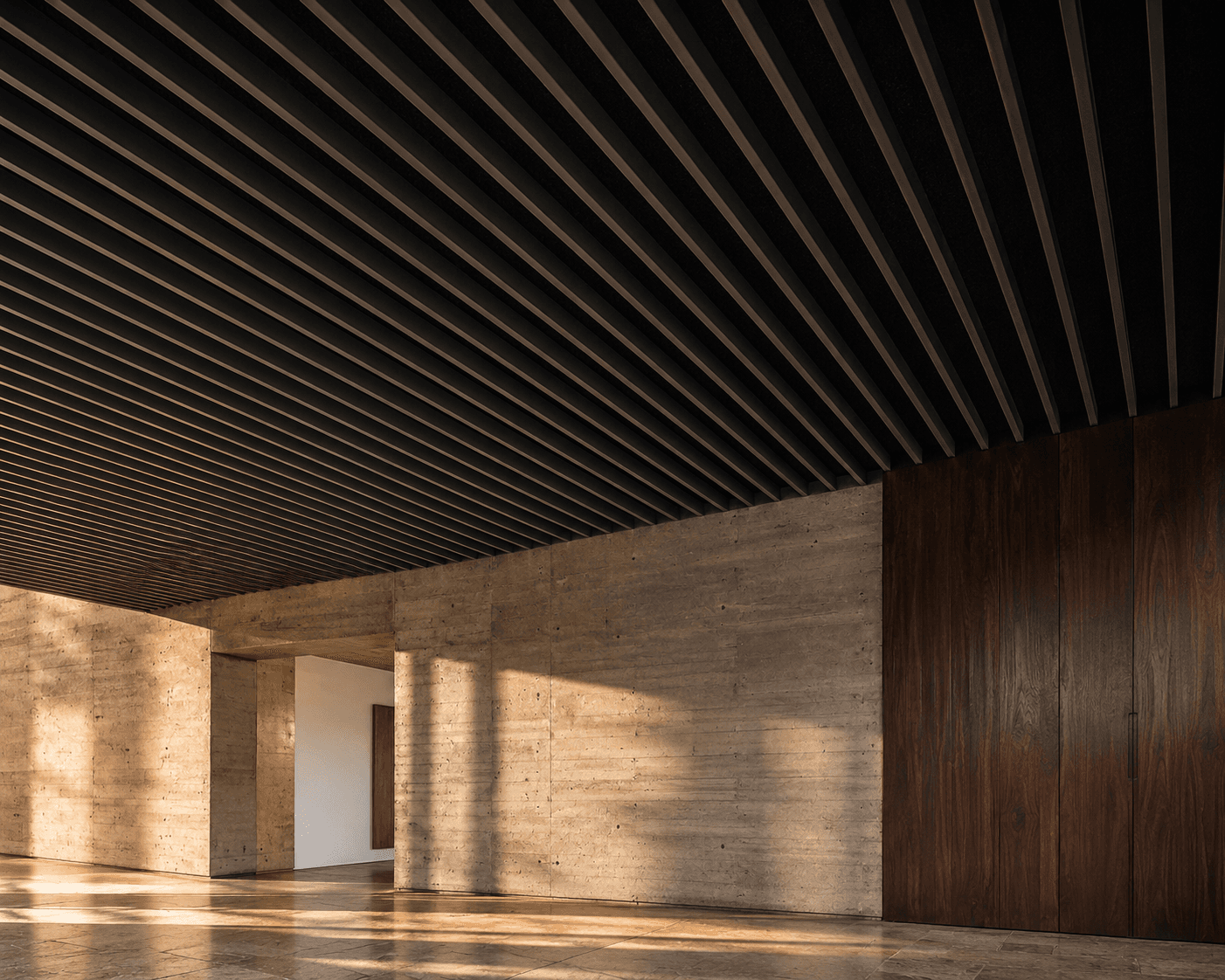 Modern interior featuring a dark linear metal baffle ceiling, board-formed concrete walls, dark wood paneling, and sunlight casting shadows across the stone floor.