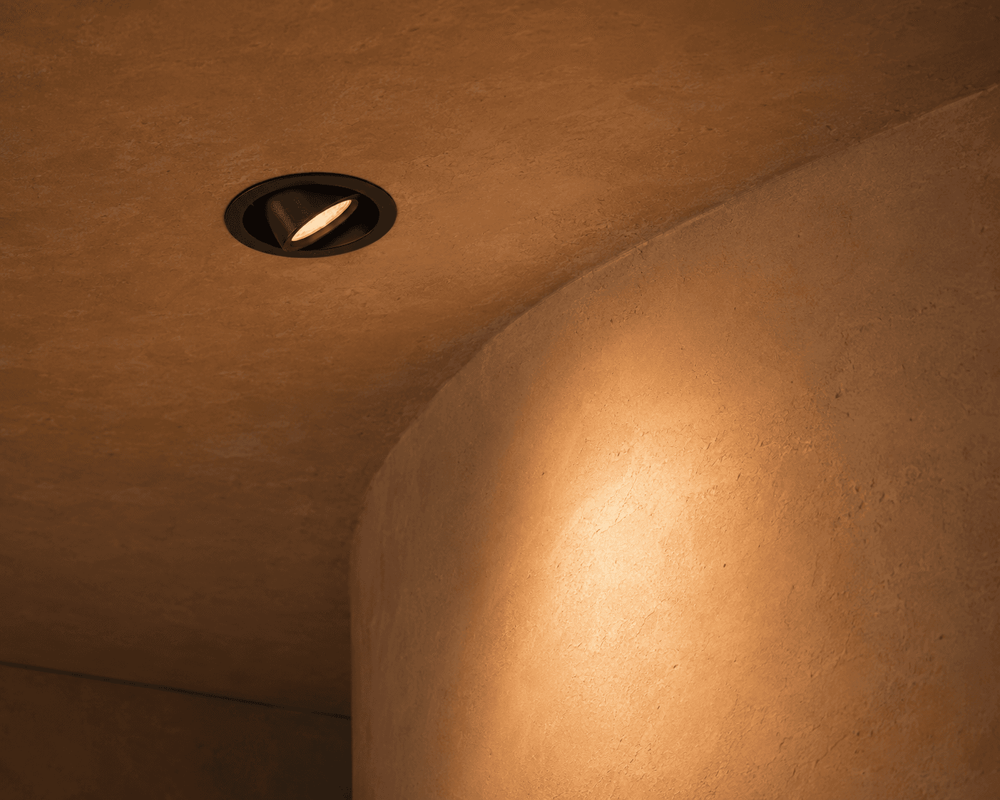 Textured beige ceiling and curved wall corner illuminated by a black recessed adjustable downlight.